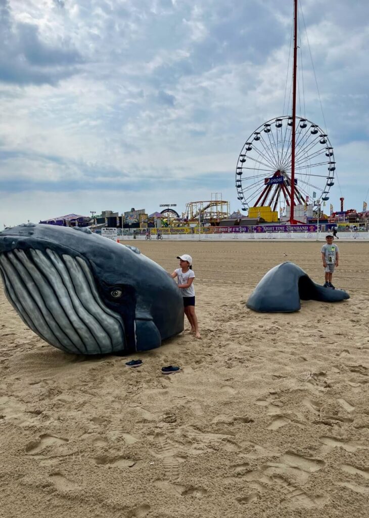 Kids playing on a whale sculpture on the beach of Ocean City, Maryland. Amusement park rides in the background.