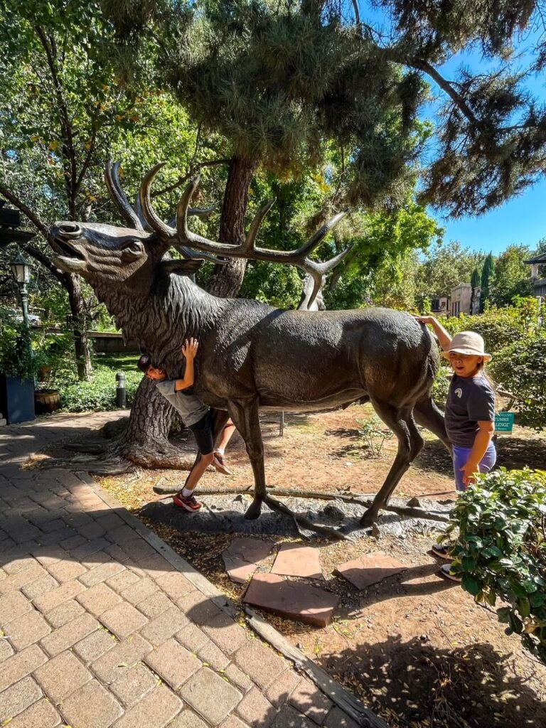 A sculpture of a moose made out of metal, 2 kids posing with it. 