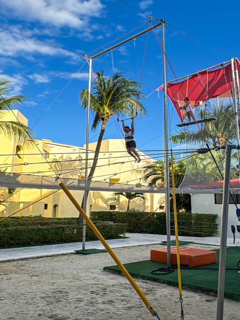 A picture of a women swinging from a trapeze bar high in the air with the hotel in the background on a sunny blue sky day.