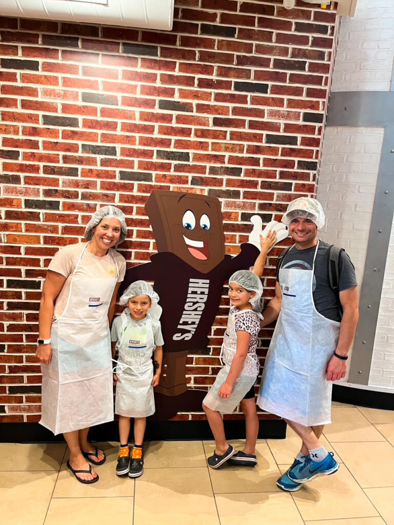 Family of 4 dressed in chef's hats and aprons posing with a picture of a Hershey Chocolate character. 