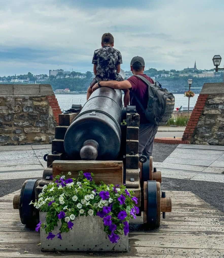 father standing beside boy as he sits on a large cannon overlooking the St. Lawrence River in Quebec City.