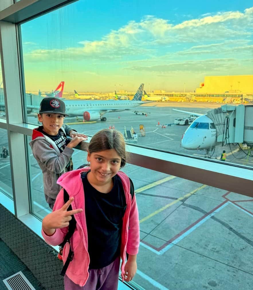 Girl and boy standing at a large window in the airport with 2 planes in the background